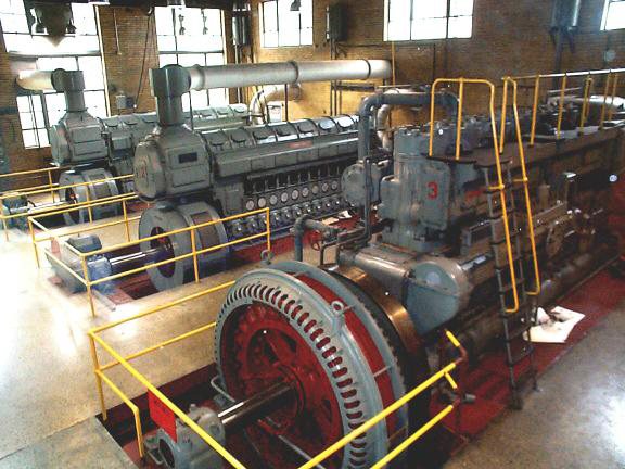 Row of large generator engines with red housings and yellow safety railings inside the power plant.