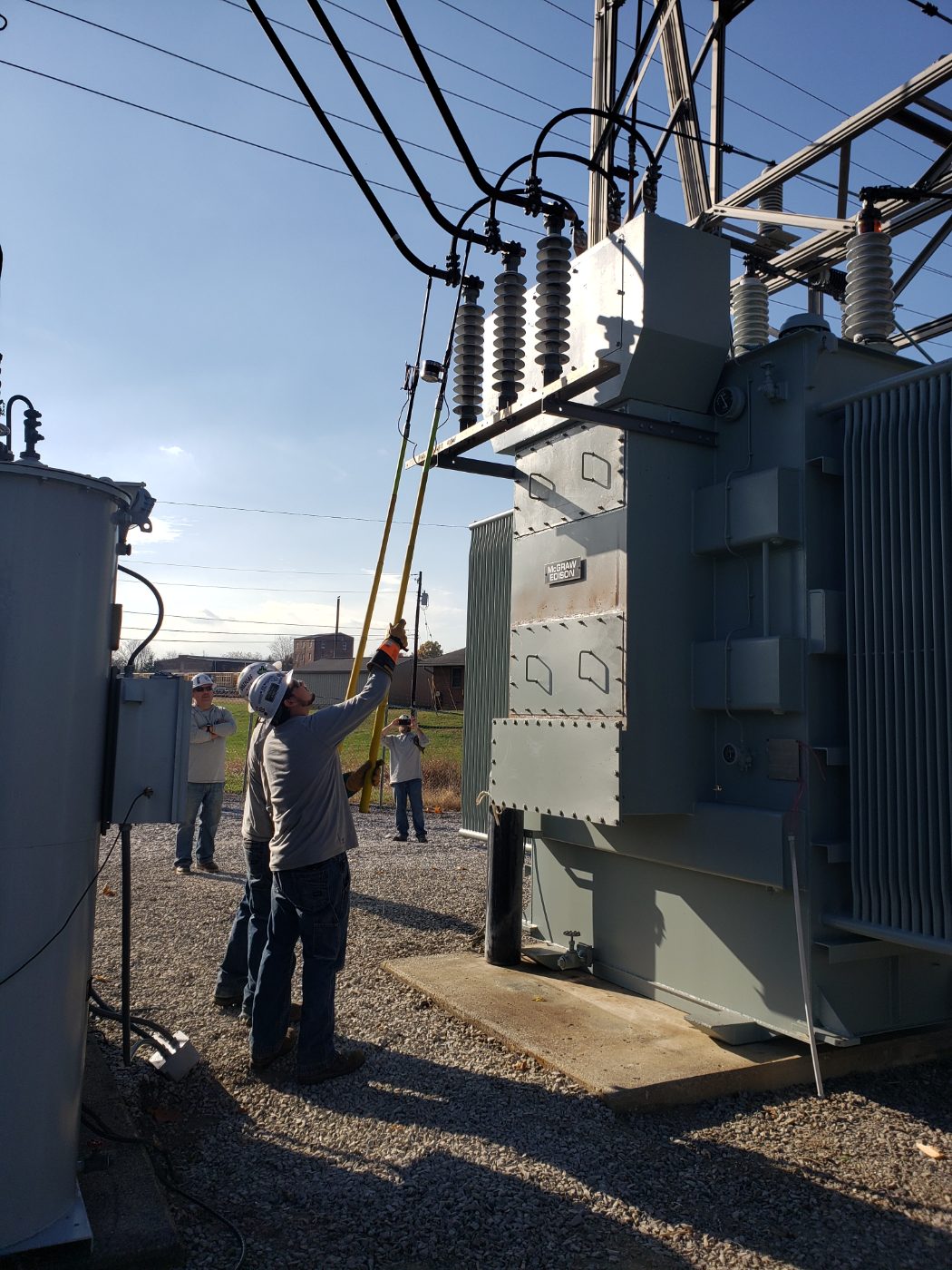 Line workers in hard hats working beside large electrical equipment on a gravel site.