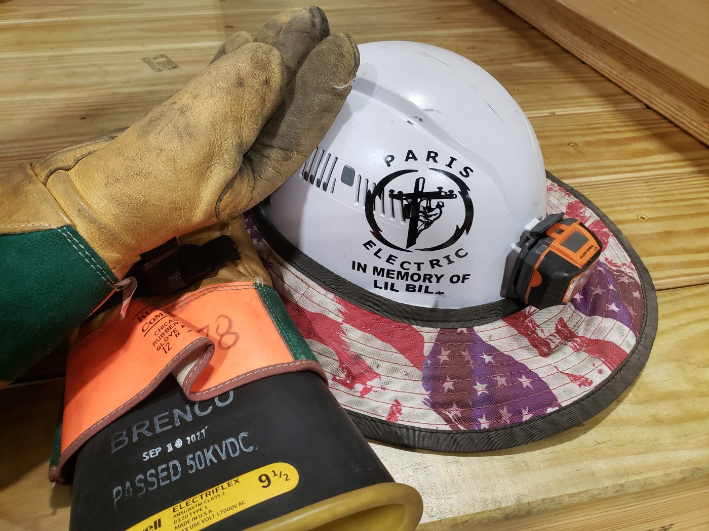 A Paris Electric hard hat reading In Memory of Lil Bill, resting beside a lineman's work gloves.