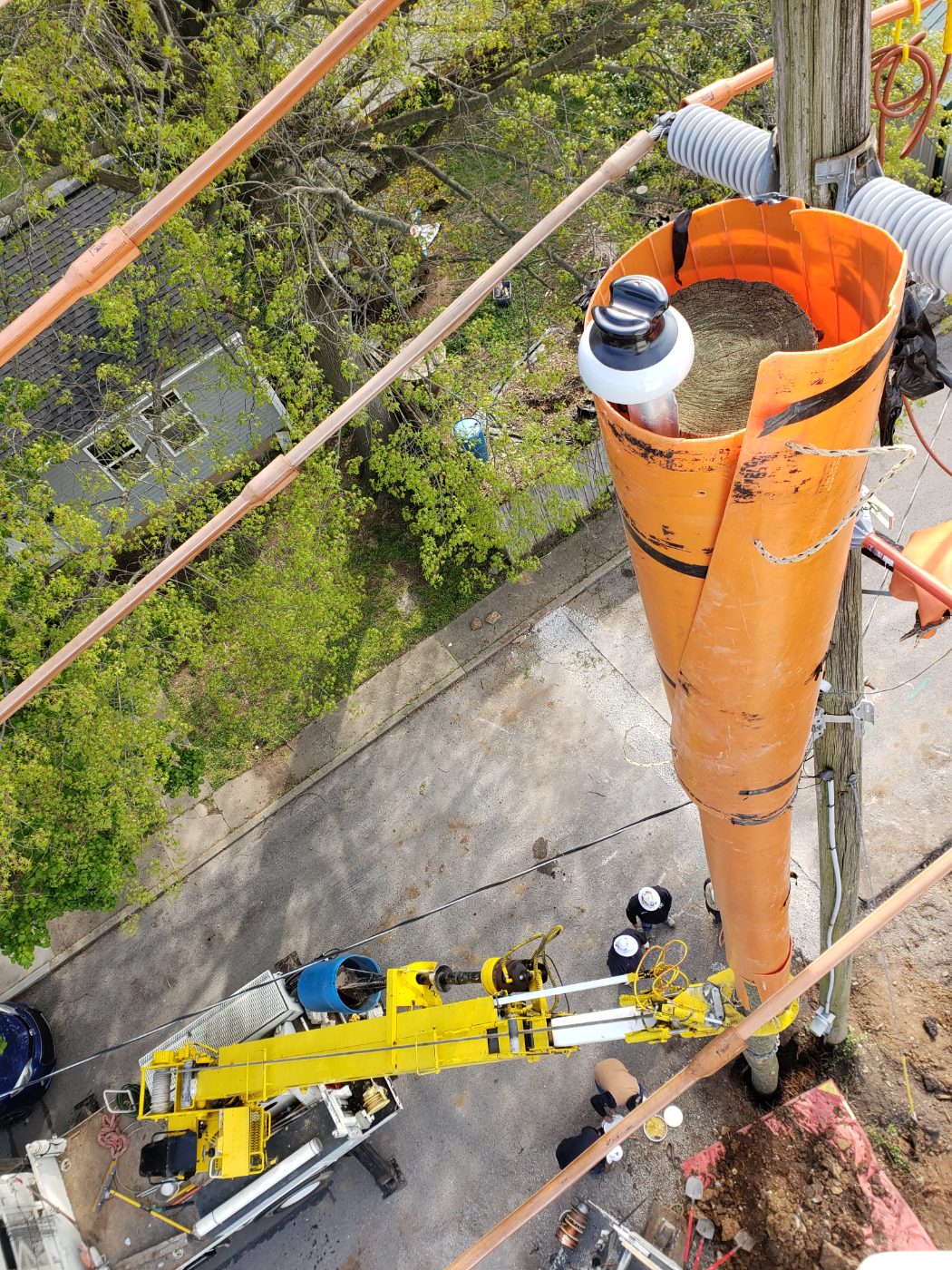 View from a bucket truck looking down a utility pole wrapped in orange protective cover, with a truck boom below.