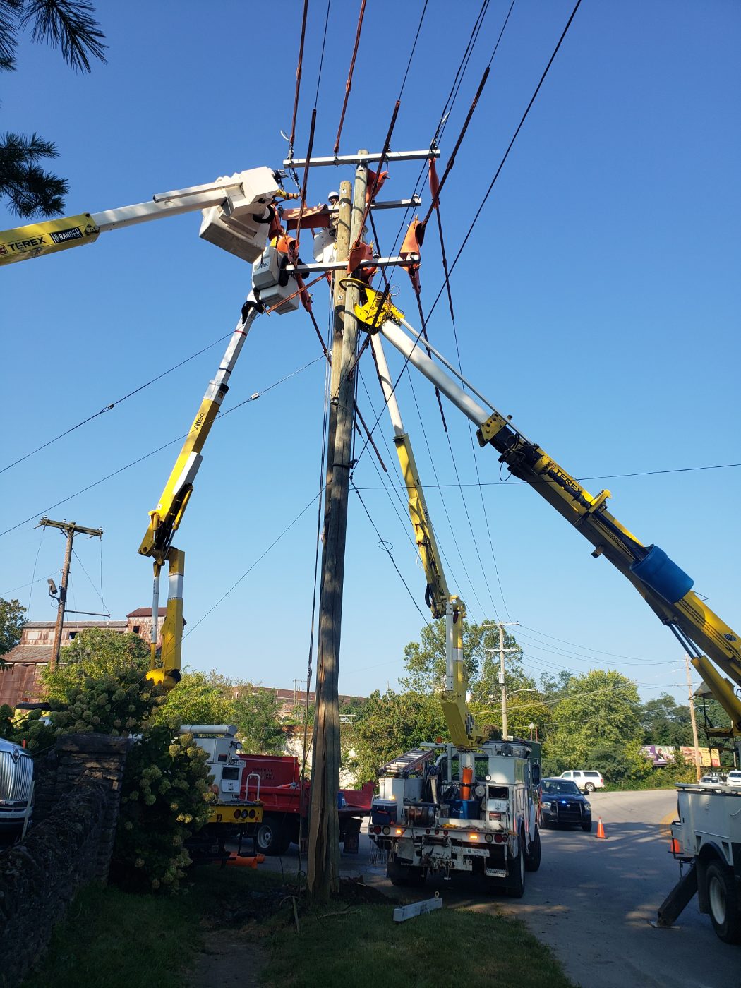 Three bucket trucks with line workers raised around a utility pole on a sunny day.